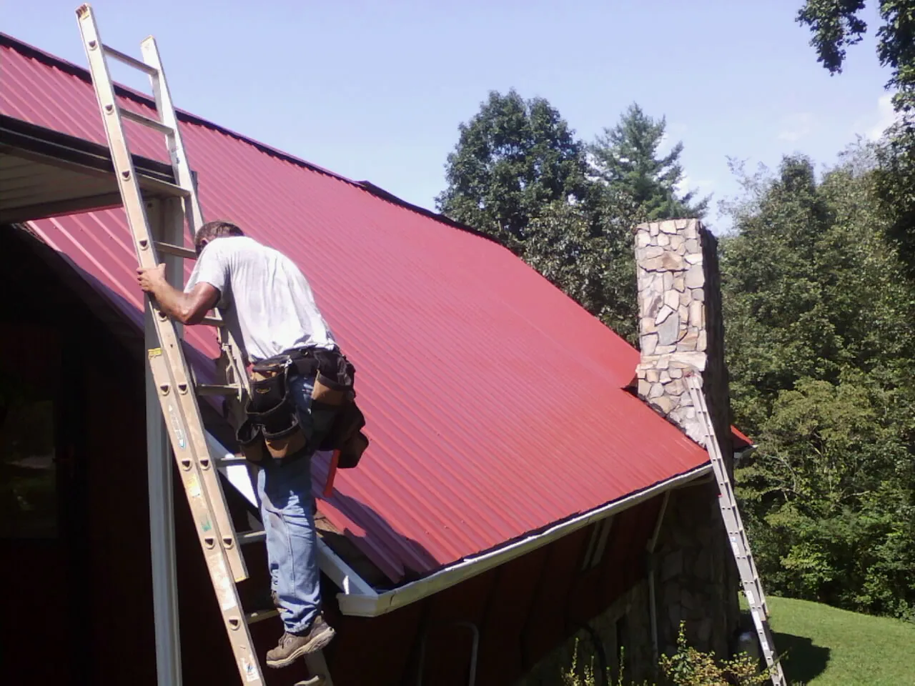 Roofer on a ladder beside a red metal roof and stone chimney — Soffit & Fascia Repair by Roofing Brothers in Canonsburg